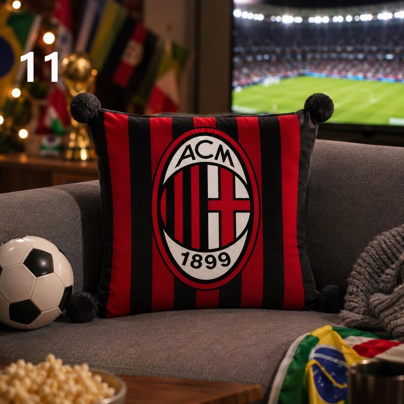 K&ouml;nnte beinhalten: Ein quadratisches Zierkissen mit dem Logo des Fu&szlig;ballclubs AC Milan in der Mitte, umgeben von vertikalen roten und schwarzen Streifen. Das Kissen hat an jeder der vier Ecken kleine schwarze Bommeln und zeigt den Text ACM 1899.