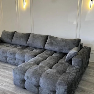 May include: A large, grey sectional sofa with a tufted design. The sofa has multiple cushions and a chaise lounge. The room has white walls and a light-coloured wood floor. Two wall sconces are mounted on the wall.