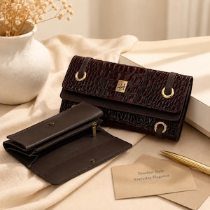 May include: A dark brown wallet with a crocodile-embossed texture, featuring gold-tone metal hardware, a central logo plaque, and a matching open wallet beside it. A small card nearby displays the text: Timeless Style Everyday Elegance.