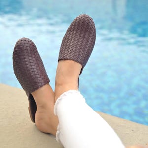 May include: A pair of dark brown woven leather mules with a closed, rounded toe design. The footwear features a detailed basket-weave pattern across the entire upper surface, providing a textured appearance and a slip-on style for easy wear.