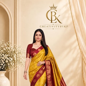 Op de afbeelding: Een mosterdgele sari met een dieprode rand met ingewikkelde gouden geweven patronen. De bijpassende bordeauxrode blouse heeft korte mouwen en een hartvormige halslijn. De stof heeft een subtiele glans en is versierd met kleine gouden motieven.