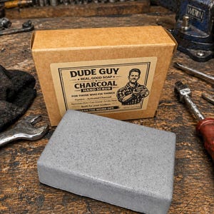 May include: A gray charcoal hand scrub soap bar next to its brown cardboard box. The box has a label with the text "DUDE GUY CHARCOAL HAND SCRUB" and a drawing of a man. The soap is for those who fix things.