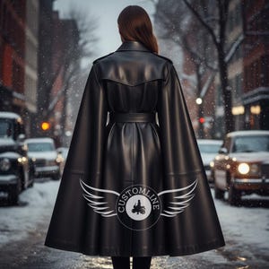 Op de afbeelding: Een zwarte leren trenchcoat met een cape-stijl rug, een ceintuur in de taille en epauletten op de schouders. De rug toont een cirkelvormig logo met de tekst "CUSTOMLINE" en een motorgrafiek, geflankeerd door gestileerde witte vleugels onderaan.