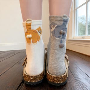 Può includere: Un paio di calzini originali, uno bianco con un disegno di gatto arancione e l'altro grigio con un disegno di gatto blu. Entrambi i calzini presentano il posteriore di un gatto con dettagli a pompon. I calzini sono indossati con sandali.