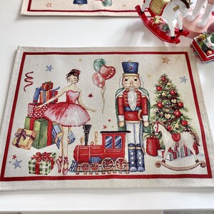 Op de afbeelding: Rechthoekige placemat met een bedrukte kerstscène met een ballerina in een roze tutu, een notenkraker-soldaat, een rode speelgoedtrein, een versierde kerstboom, een hobbelpaard en gestapelde cadeaus, omlijst door een effen rode rand.
