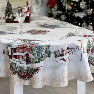 Puede incluir: Un mantel ovalado con una escena invernal de casas rojas, pinos nevados, muñecos de nieve y vallas. El diseño incluye bordes decorativos con campanas y copos de nieve sobre un fondo blanco, cubriendo una mesa de comedor con vajilla.