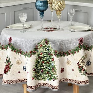 Op de afbeelding: Een rond tafelkleed met een feestelijke kerstboomprint met hangende ornamenten, groene slingers en rode kerststerren op een crèmekleurige achtergrond met een zilveren rand. De tafel is gedekt met twee witte borden en vier decoratieve glazen.