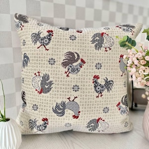 May include: Square cushion featuring a cream background with a repeating pattern of roosters in grey, black, and red. The cushion has a geometric design and is a farmhouse style.