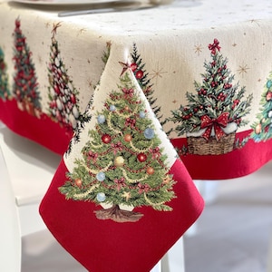 Könnte beinhalten: Eine cremefarbene Tischdecke mit einem sich wiederholenden Muster aus geschmückten Weihnachtsbäumen mit roten, goldenen und blauen Ornamenten, goldenen Sternen und einem einfarbigen roten Rand am unteren Saum. Der Stoff hat eine Webstruktur.