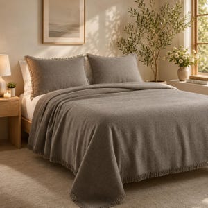 May include: A solid taupe-colored bedspread and two matching pillow shams, each featuring a textured fabric finish and decorative fringe detailing along the edges. The bedding set is draped over a bed, showcasing a consistent neutral tone throughout.