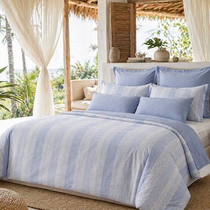 May include: A bed with a light blue and white striped duvet cover and matching pillowcases. The bedding is arranged on a white bed frame, with a wooden side table and a woven basket with a plant in the background. The room has a bright, airy feel.