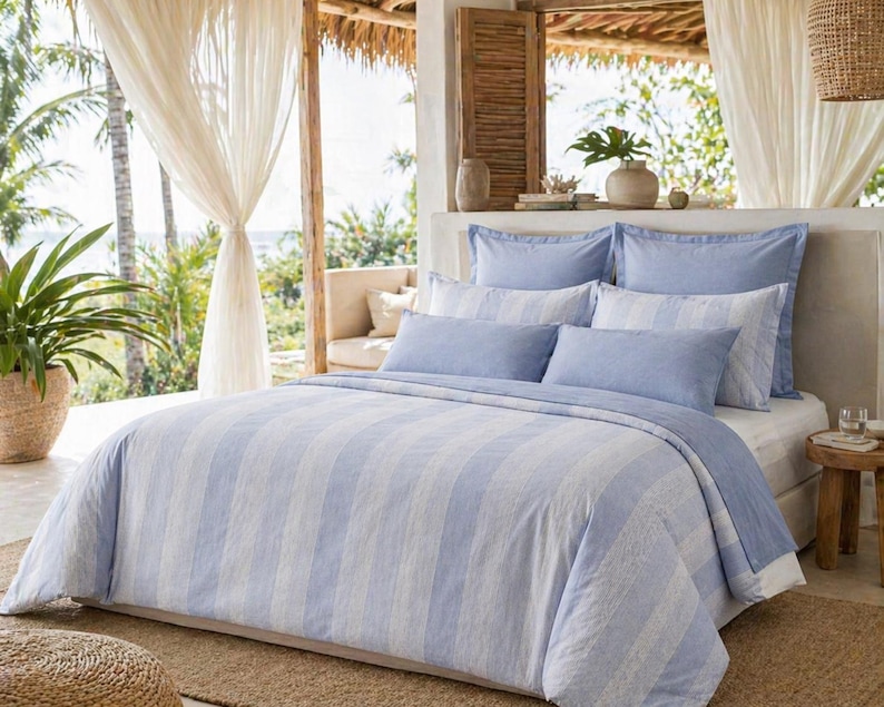 May include: A bed with a light blue and white striped duvet cover and matching pillowcases. The bedding is arranged on a white bed frame, with a wooden side table and a woven basket with a plant in the background. The room has a bright, airy feel.