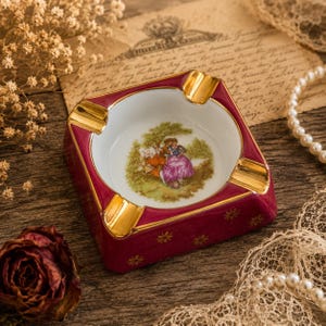 May include: A square porcelain ashtray featuring a deep burgundy exterior with gold floral accents and four gold-rimmed notches. The center of the white basin displays a colorful illustration of two figures sitting under a tree in a landscape.