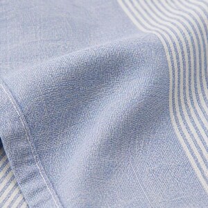 May include: Close-up of a light blue and white striped fabric. The fabric has a textured appearance with visible woven threads. The stripes are vertical and alternate between light blue and white. The fabric is folded, showing the texture and the stitching.