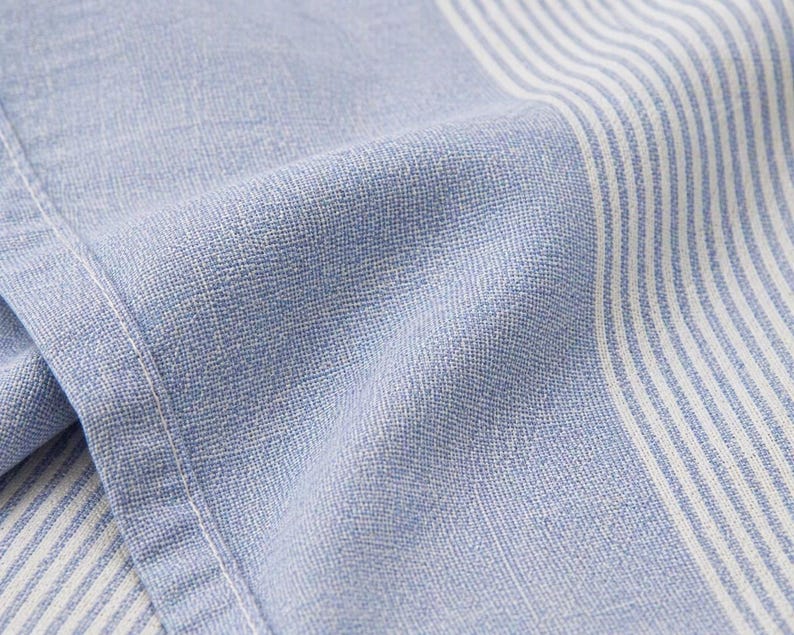 May include: Close-up of a light blue and white striped fabric. The fabric has a textured appearance with visible woven threads. The stripes are vertical and alternate between light blue and white. The fabric is folded, showing the texture and the stitching.