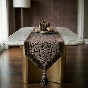 May include: A marble-topped table with a decorative runner and gold accents. The runner is brown with an ornate pattern and a tassel. Gold candle holders and decorative stones sit on the table.