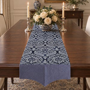 May include: A rectangular table runner featuring a dark blue and light blue damask pattern with a solid light blue quilted border. The runner is centered on a dark wood dining table, positioned beneath a floral centerpiece and two silver candlesticks.