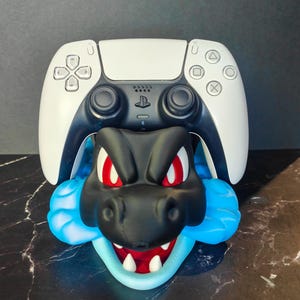 May include: A white and black video game controller resting on a black and blue character stand. The stand features a stylized black creature head with red eyes, a wide open mouth with white teeth, and bright blue cloud-like accents on the sides.