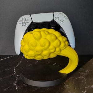 May include: A white and black video game controller rests on a yellow, cloud-shaped stand. The stand features a textured, rounded yellow cloud element attached to a solid black circular base, providing a stable mount for the gaming peripheral.