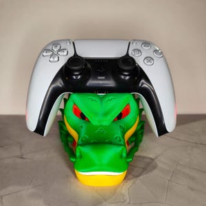 Peut inclure: Une manette blanche et noire PlayStation 5 repose sur un support en forme de dragon vert, jaune et rouge. Le dragon a des yeux rouges et un design texturé détaillé. La manette est centrée au-dessus du dragon.