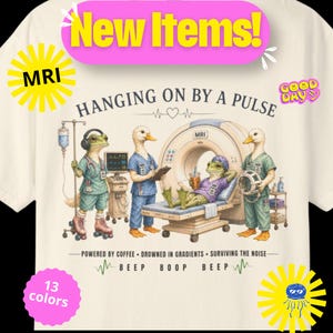 May include: A cream-colored t-shirt featuring a graphic of frogs and ducks in medical scrubs around an MRI machine. Text reads: HANGING ON BY A PULSE, POWERED BY COFFEE, DROWNED IN GRADIENTS, SURVIVING THE NOISE, BEEP BOOP BEEP, NEW ITEMS, MRI, GOOD DAY, 13 colors.