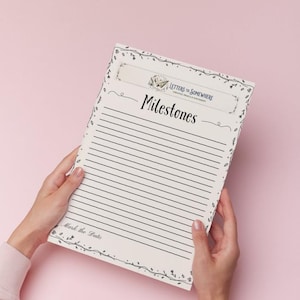 May include: A cream-colored milestone journal with lined pages, held against a pink background. The journal features the text "Milestones" in elegant script, with decorative floral accents and the phrase "Letters to Somewhere" at the top.