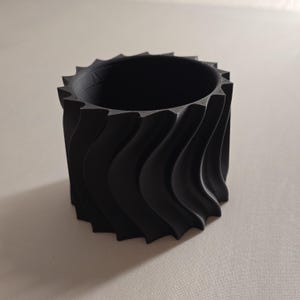 Könnte beinhalten: Ein schwarzer zylindrischer Behälter mit einer gewellten, strukturierten Außenseite. Der obere Rand weist eine Reihe von dreieckigen Spitzen auf. Der Innenraum ist hohl. Das Design ist modern und geometrisch. Das Objekt scheint aus einem festen Material zu bestehen.