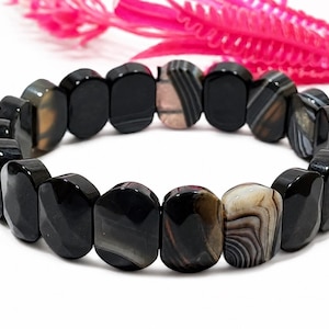 Könnte beinhalten: Ein elastisches Armband bestehend aus facettierten, ovalen Perlen in den Farben Schwarz, Dunkelbraun und Grau. Die Perlen weisen natürliche Streifenmuster sowie glatte, polierte Oberflächen auf und sind in einer kreisförmigen Schlaufe angeordnet.