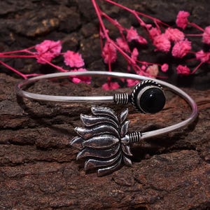 Könnte beinhalten: Silberfarbenes Armreif-Armband mit einem schwarzen Edelstein und einem detaillierten Lotusblumen-Design. Das Armband hat ein gebogenes, offenes Design und liegt vor einem strukturierten, braunen Hintergrund mit rosa floralen Akzenten.