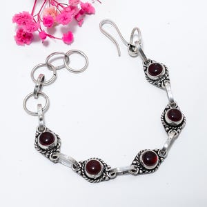 May include: A silver bracelet featuring oval, dark red gemstones set in decorative bezels. The bracelet has a chain-link design with a hook clasp. Pink flowers are in the upper left corner.