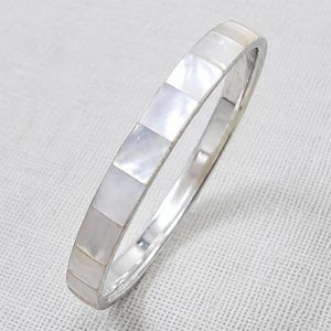 May include: A silver-toned bangle bracelet featuring a series of inlaid, square-shaped mother-of-pearl tiles. The bracelet has a smooth, polished metal frame that surrounds the iridescent, white, and cream-coloured shell segments in a continuous row.