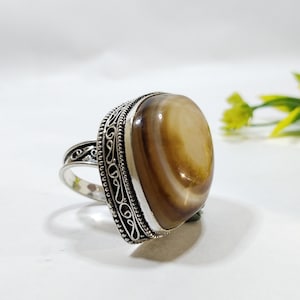 May include: A silver ring featuring a large, smooth, oval-shaped stone with concentric bands of brown, tan, and cream colors. The metal setting displays intricate filigree scrollwork and a beaded border surrounding the central gemstone.