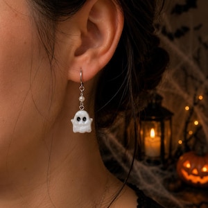 May include: A silver-toned drop earring featuring a small, white, glossy ghost charm with two black oval eyes. The ghost is suspended from a silver metal hoop by a thin chain accented with a single small, round, white pearl bead.