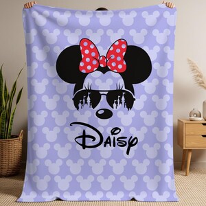 May include: A light purple blanket featuring a large Minnie Mouse graphic with sunglasses and a red polka dot bow. The blanket has a repeating pattern of white Mickey Mouse heads and the name "Daisy" in black.