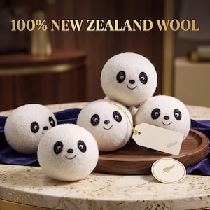 May include: Five white felted wool balls shaped like panda faces with black eyes and small smiles. The text 100% NEW ZEALAND WOOL is displayed above. A cream-colored rectangular tag with a fern icon and a matching round tag are attached to the balls.
