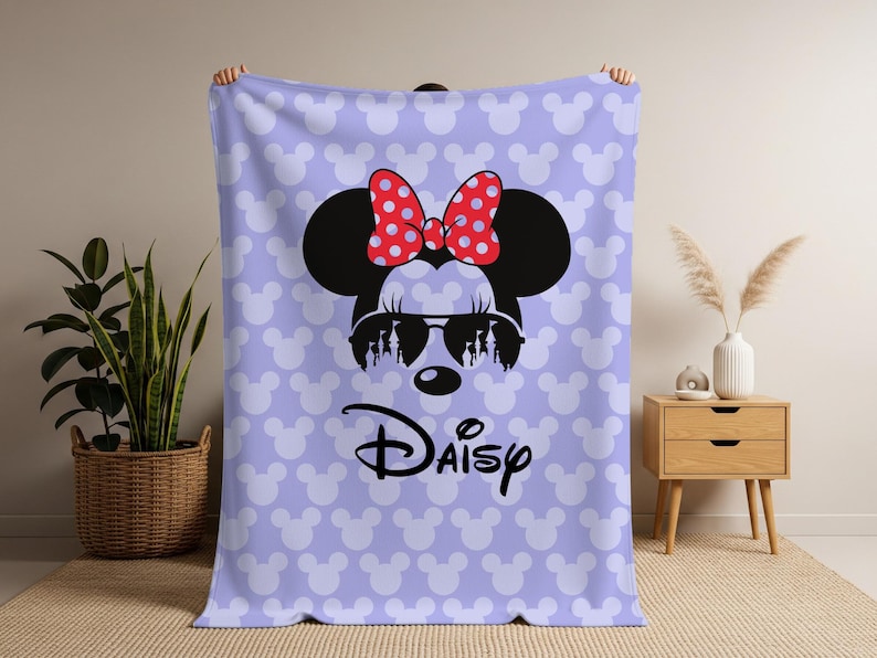 May include: A light purple blanket featuring a large Minnie Mouse graphic with sunglasses and a red polka dot bow. The blanket has a repeating pattern of white Mickey Mouse heads and the name "Daisy" in black.