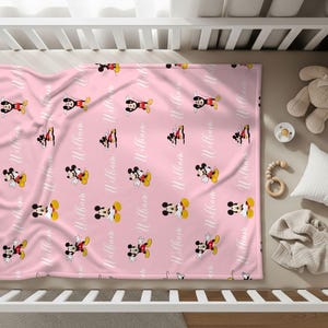 Puede incluir: Manta rosa para beb&eacute; con un patr&oacute;n repetido de Mickey Mouse en rojo, amarillo, negro y blanco. El nombre "William" est&aacute; impreso en escritura blanca. La manta est&aacute; en una cuna con un osito de peluche, un chupete y una almohada.