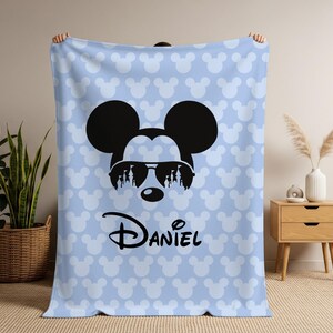 May include: A light blue blanket featuring a repeating pattern of white Mickey Mouse heads. The blanket has a large black Mickey Mouse head wearing sunglasses with a silhouette design. The name "Daniel" is printed in black.
