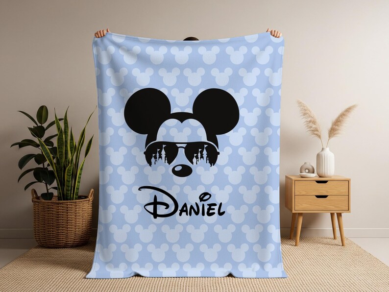 May include: A light blue blanket featuring a repeating pattern of white Mickey Mouse heads. The blanket has a large black Mickey Mouse head wearing sunglasses with a silhouette design. The name "Daniel" is printed in black.
