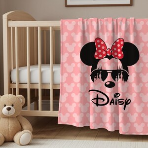 May include: Pink baby blanket featuring a Minnie Mouse design. The blanket has a pattern of white Mickey Mouse heads and a large Minnie Mouse graphic with sunglasses and a red polka dot bow. The word "Daisy" is written below the graphic.