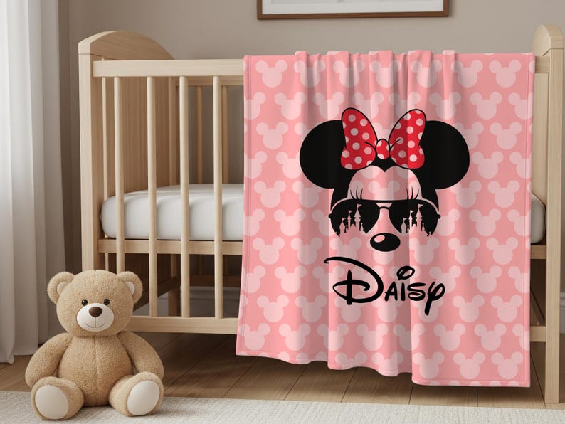 May include: Pink baby blanket featuring a Minnie Mouse design. The blanket has a pattern of white Mickey Mouse heads and a large Minnie Mouse graphic with sunglasses and a red polka dot bow. The word "Daisy" is written below the graphic.