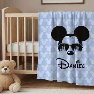May include: A light blue baby blanket with a Mickey Mouse design. The blanket features a large black Mickey Mouse head wearing sunglasses, with the name "Daniel" in script. The background has a pattern of white Mickey Mouse heads.