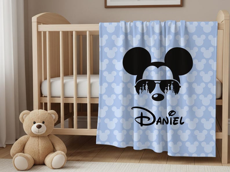 May include: A light blue baby blanket with a Mickey Mouse design. The blanket features a large black Mickey Mouse head wearing sunglasses, with the name "Daniel" in script. The background has a pattern of white Mickey Mouse heads.