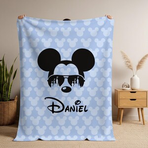 May include: A light blue blanket featuring a repeating pattern of white Mickey Mouse heads. The blanket has a large black Mickey Mouse head with sunglasses and the name "Daniel" in black script.