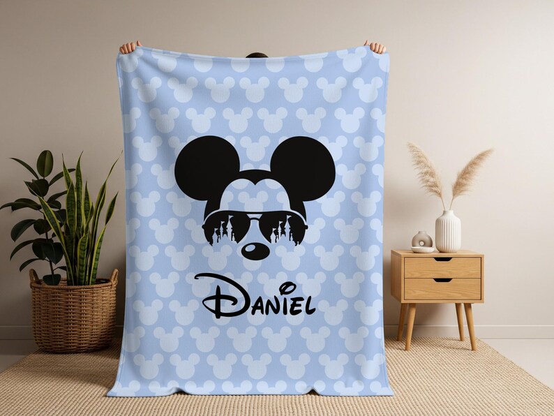 May include: A light blue blanket featuring a repeating pattern of white Mickey Mouse heads. The blanket has a large black Mickey Mouse head with sunglasses and the name "Daniel" in black script.