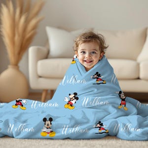 Puede incluir: Una manta de beb&eacute; azul claro con personajes de dibujos animados de Mickey Mouse y el nombre "William" en escritura blanca. La manta est&aacute; envuelta alrededor de un ni&ntilde;o sonriente. La manta es de material suave.