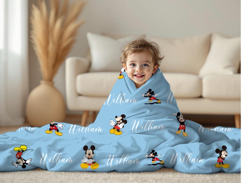Puede incluir: Una manta de beb&eacute; azul claro con personajes de dibujos animados de Mickey Mouse y el nombre "William" en escritura blanca. La manta est&aacute; envuelta alrededor de un ni&ntilde;o sonriente. La manta es de material suave.