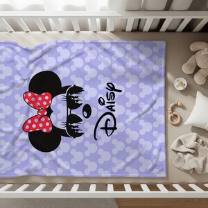 May include: A lavender baby blanket featuring a Minnie Mouse design with a red polka dot bow and the name "Daisy" in black script. The blanket is covered in white Mickey Mouse head silhouettes.
