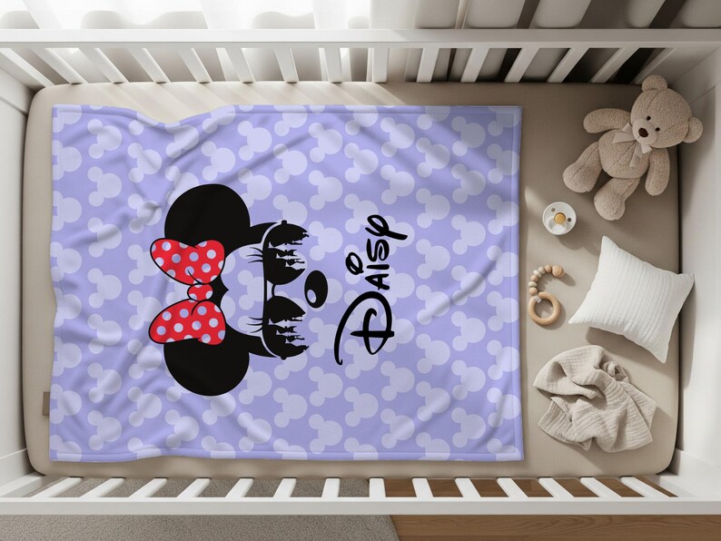 May include: A lavender baby blanket featuring a Minnie Mouse design with a red polka dot bow and the name "Daisy" in black script. The blanket is covered in white Mickey Mouse head silhouettes.