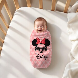 May include: A baby wrapped in a pink swaddle blanket with a Minnie Mouse design. The blanket features a pattern of small white hearts and a large Minnie Mouse graphic with sunglasses and a red bow. The word "Daisy" is written below.